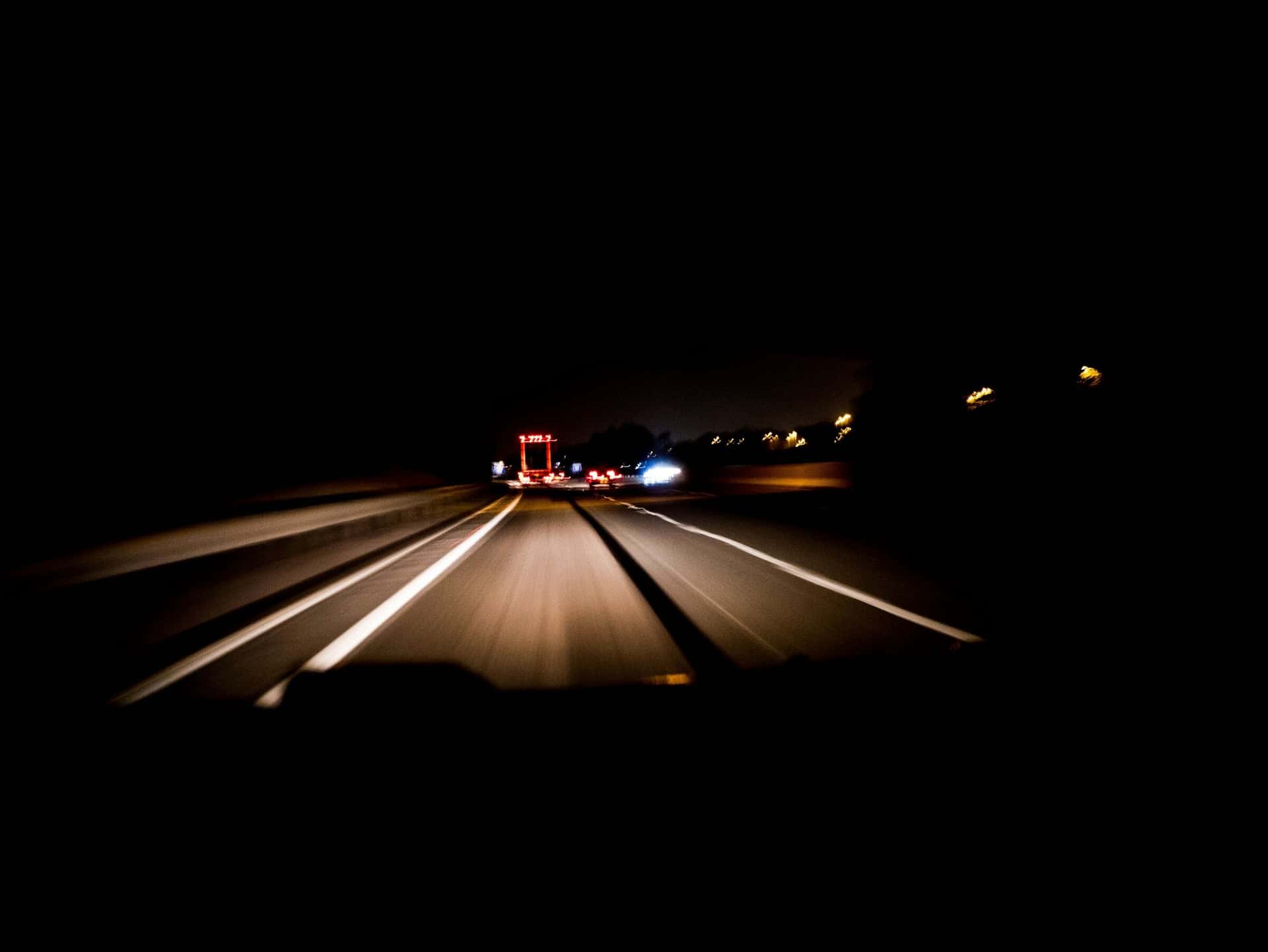 A British motorway at night, seen from a moving car, with an overhead gantry ahead.