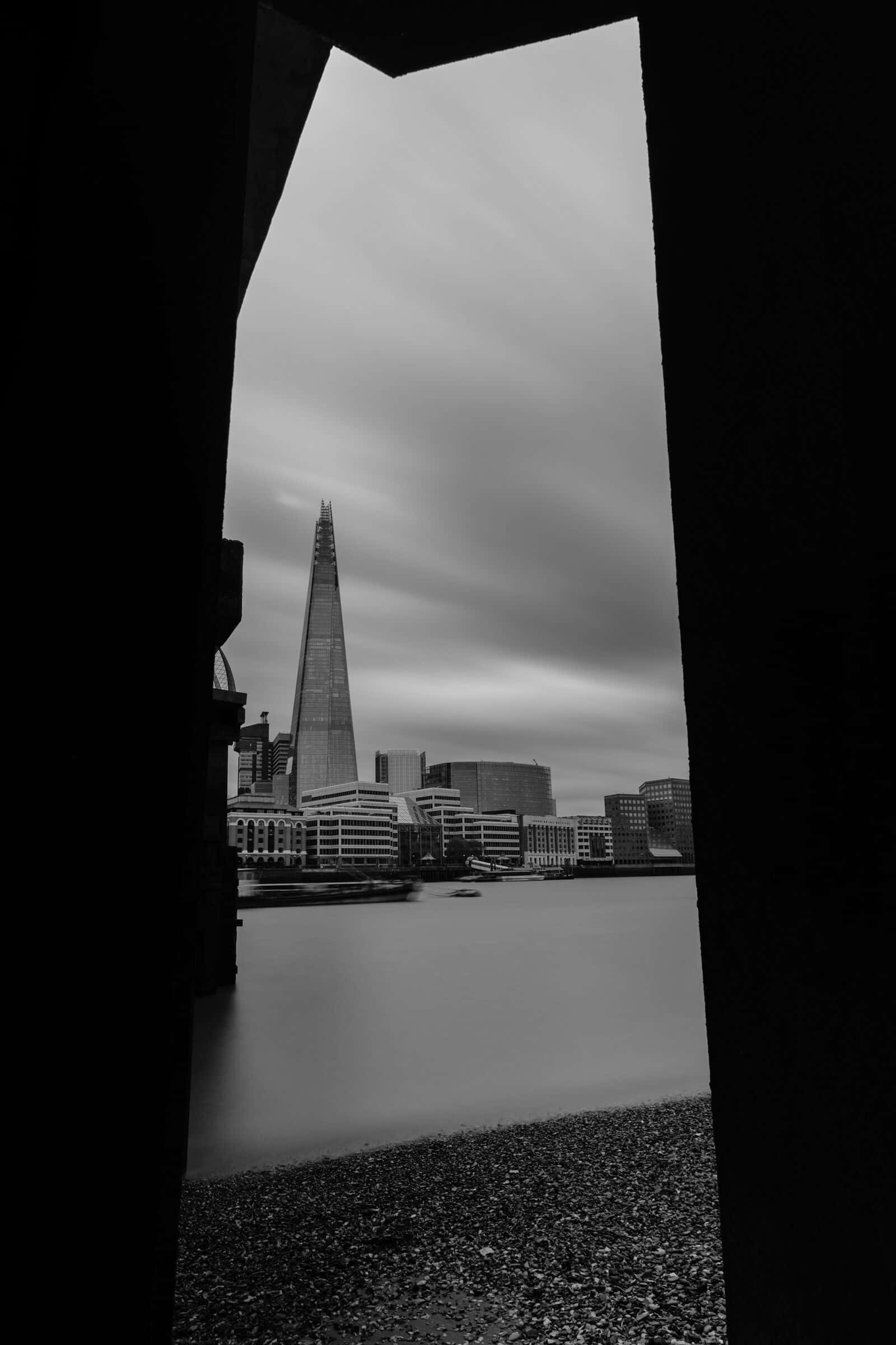The Shard seen across the Thames from the shingle foreshore, framed by the dark underside of a bridge.